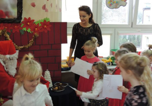 Mikołaj siedzi przy stoliku zastawionym ciasteczkami a wokół niego dzieci i nauczycielka